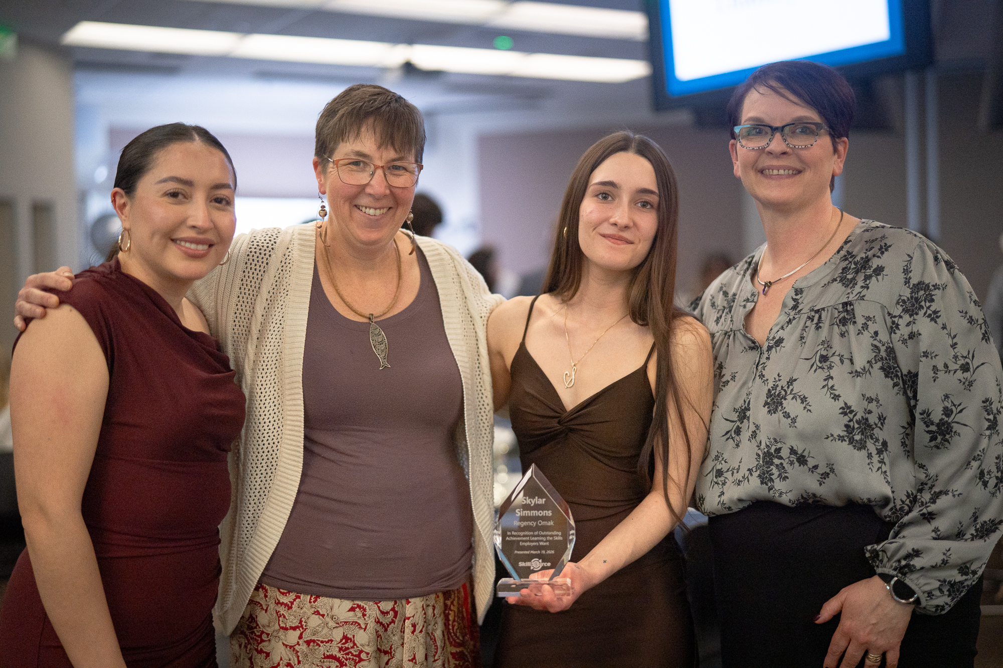 Four smiling women, diverse in age and appearance, in dressy attire, the youngest with long dark hair holds up a clear glass award.