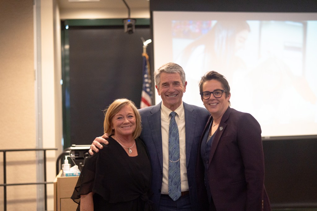 A petite blonde woman stands next to a gray-haired smiling center who stands beside a taller woman with short hair and dark glasses as they all smile in front of a projector screen and an American flag in the background.