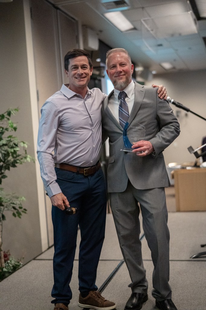 Two smiling men in collared shirts and one in a suit stand shoulder to shoulder while one holds up a clear glass award.