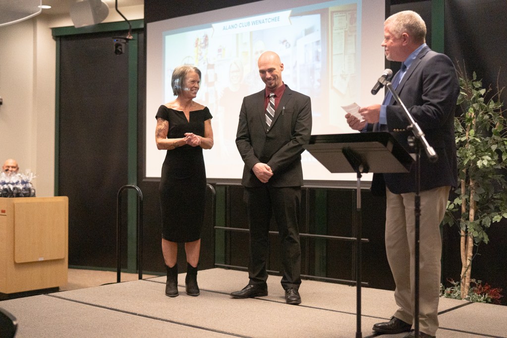 A man in a suit stands on a stage in between two other well-dressed people who applaud him.