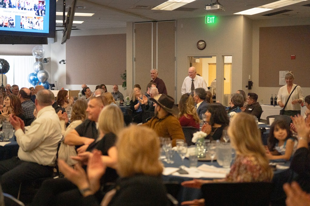 A banquet room full of people clapping as two older gentlemen at the back rise to be recognized.