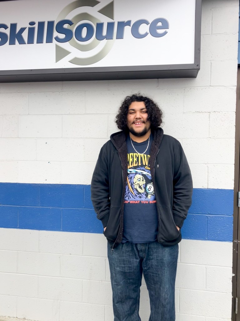 A tall man with dark hair posing for a photo in front of a wall with SkillSource's logo.