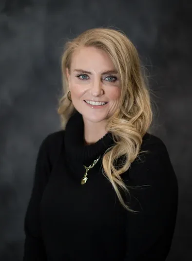 A woman with blonde hair and blue eyes wearing a black cowl neck sweater in front of a mottled gray photo backdrop.