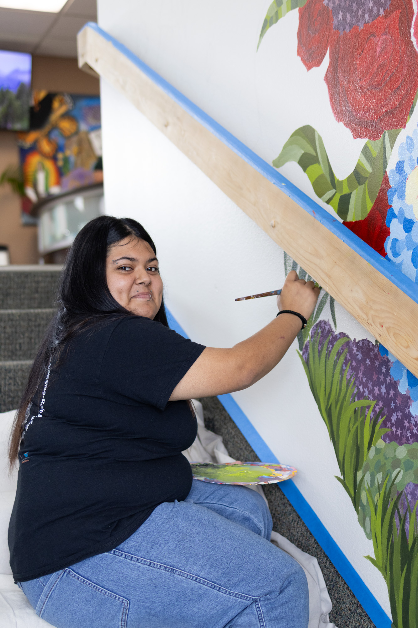 A woman wearing a black tee shirt and jeans is sitting on a set of stairs looking at the camera, holding a palette while painting a wall.