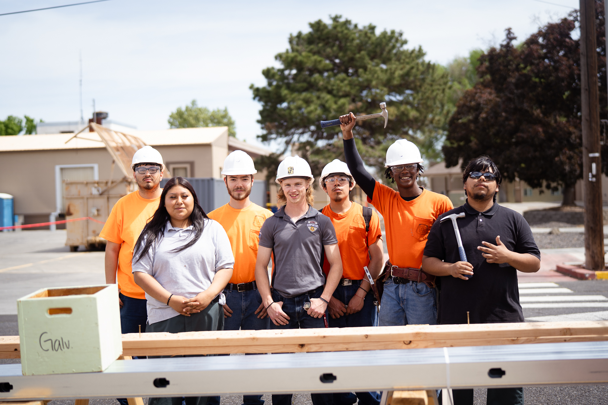 A diverse group of young people in white hard hats and orange t-shirts stand behind a carpentry demonstration table.