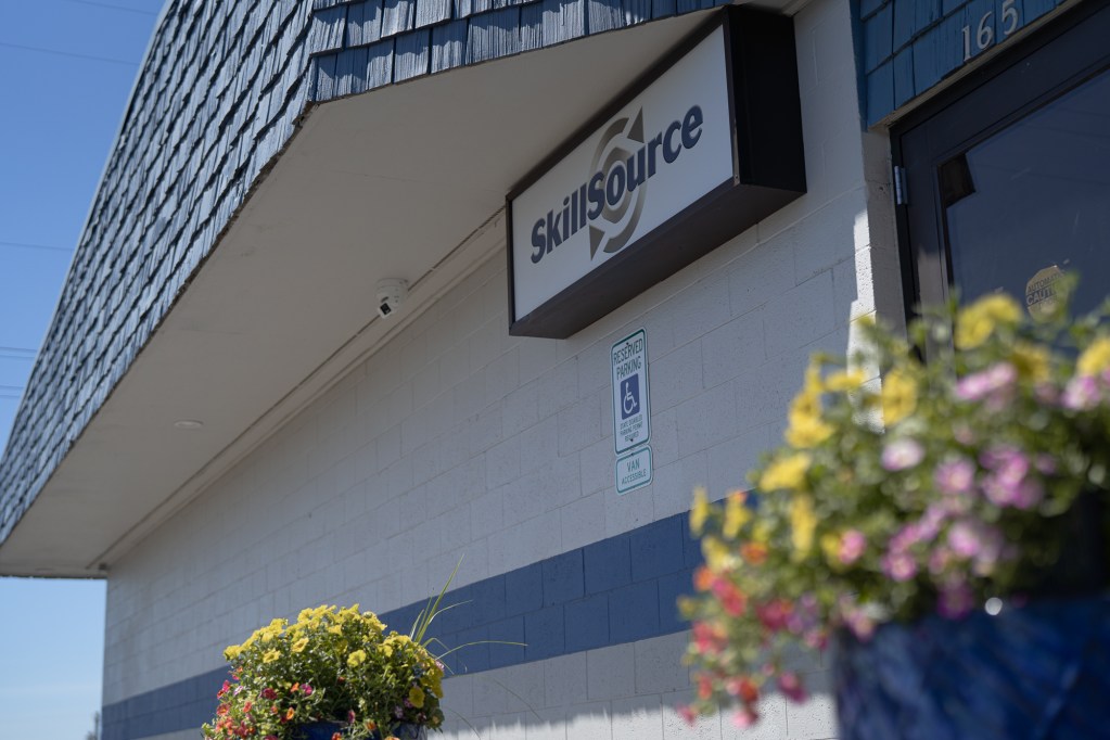 Two planters filled with bright flowers sit in front of a white building with a blue stripe and a sign that reads SkillSource.