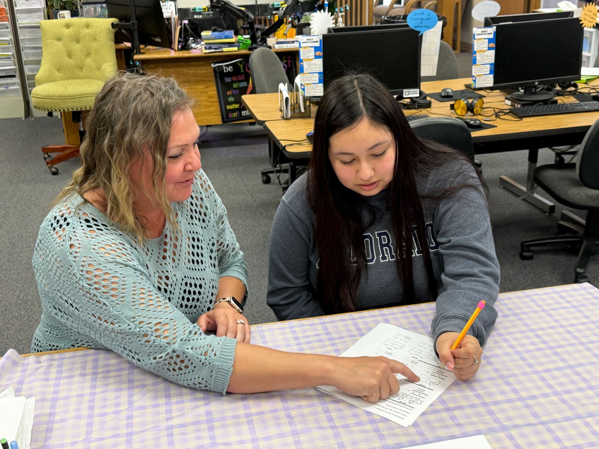 A female instructor is seated at a table with a long-haired student and they work on a sheet together.