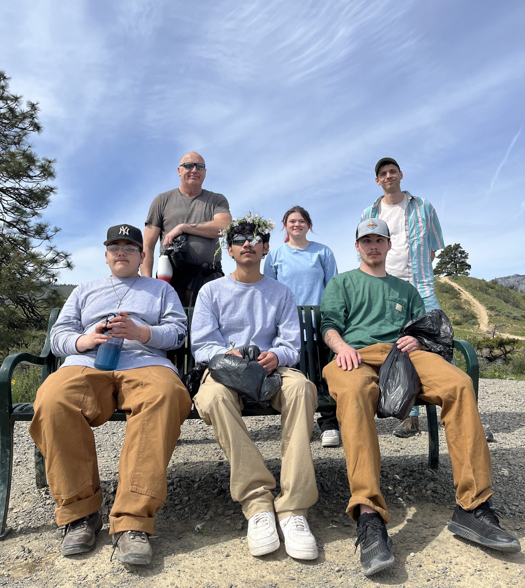 A group of four students on and behind a park bench with a bright blue sky behind them and two instructors smiling in the background.