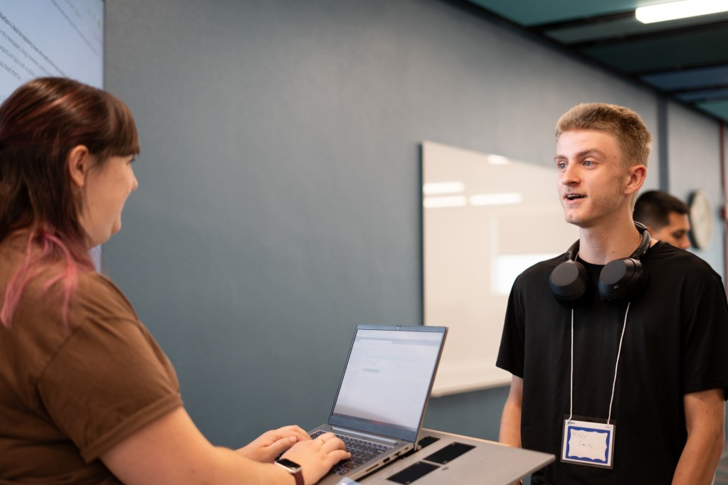 A student wearing a black t-shirt is speaking to a staff member at a library while she is on her computer.