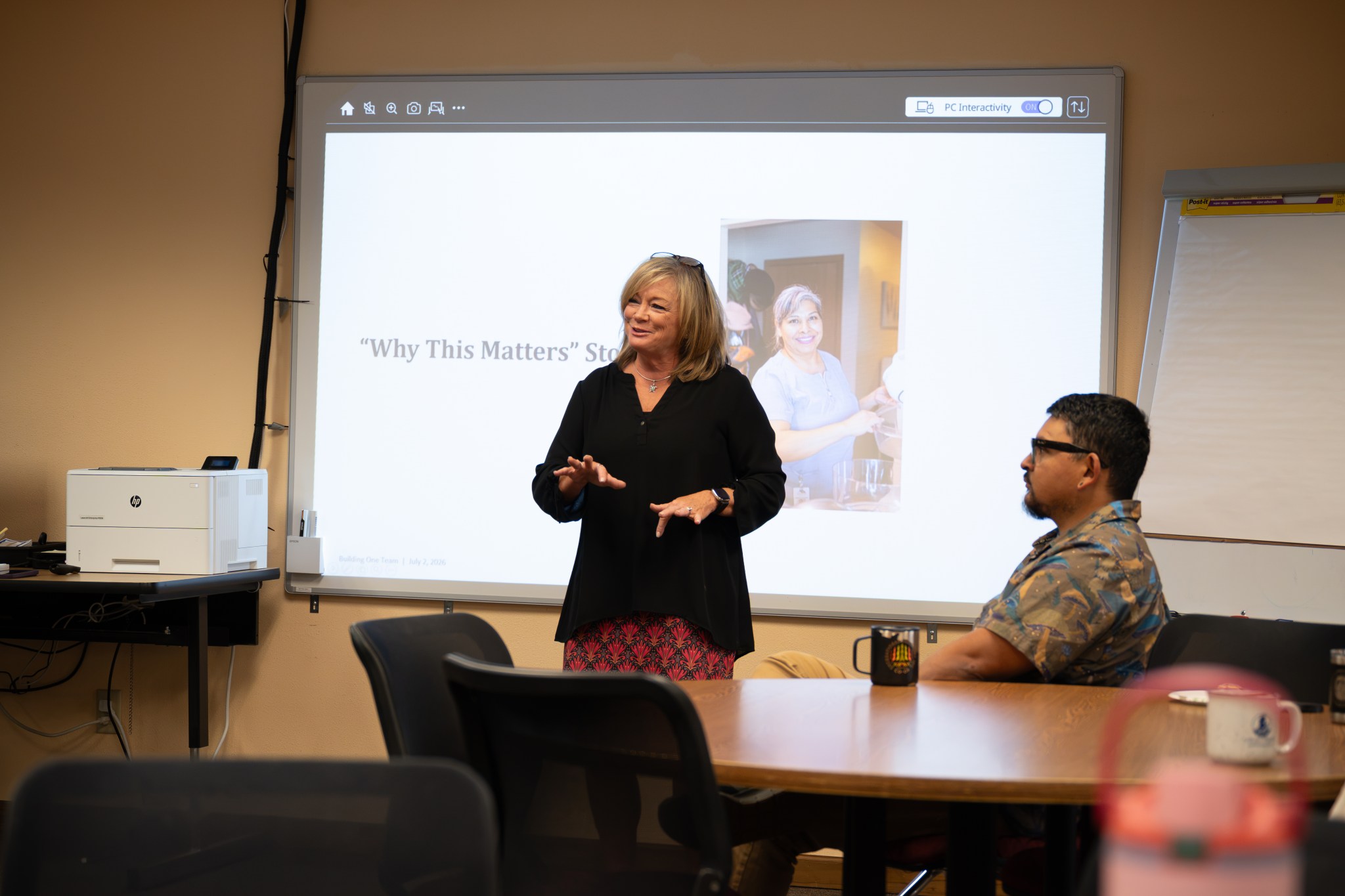 A professional woman with light hair wearing a black blouse and red skirt stands in front of a bright screen that has a photo of a woman in scrubs.