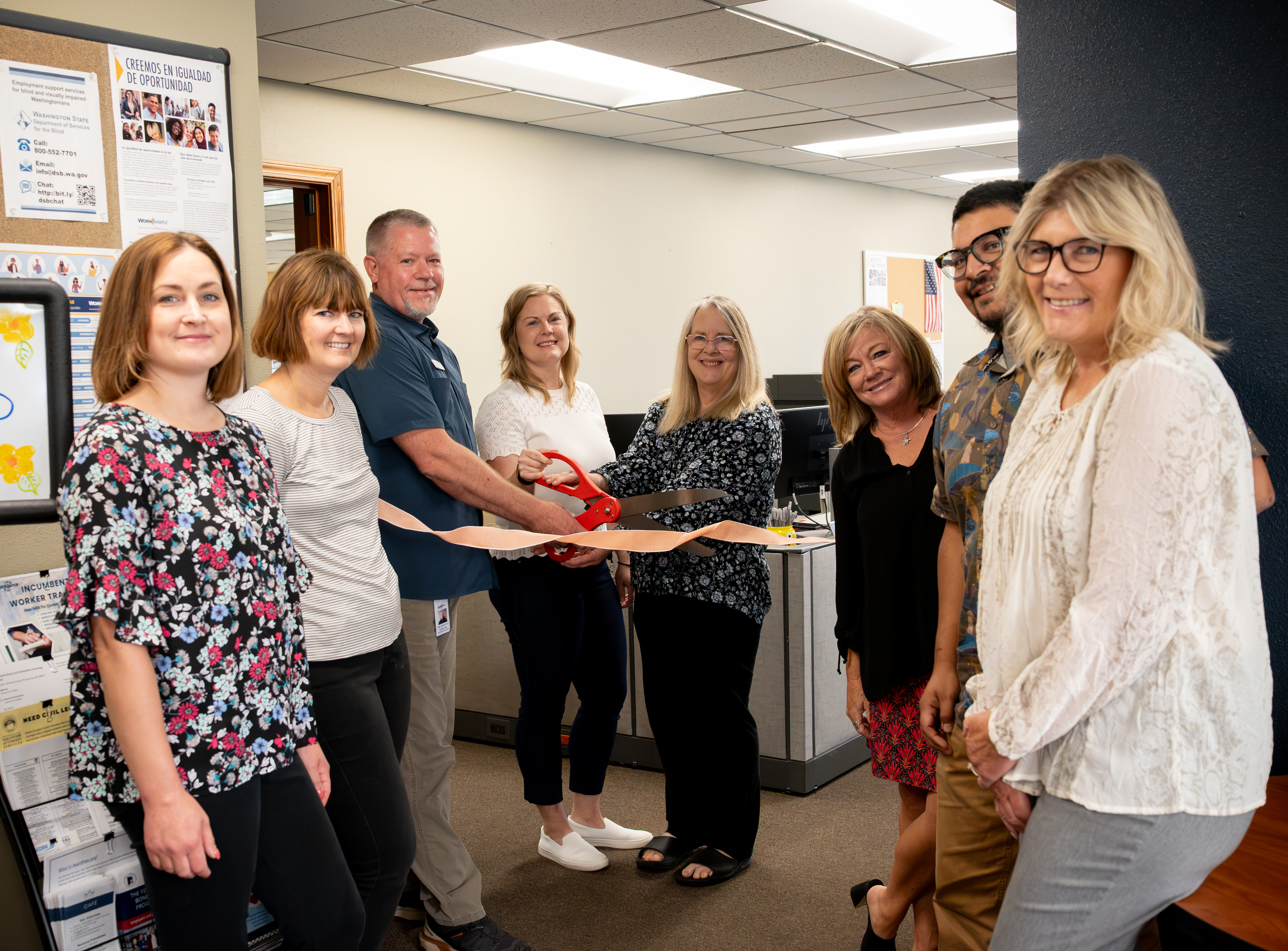 A group of 8 people stand inside an office building while a couple of them hold giant red scissors to cut a ribbon.